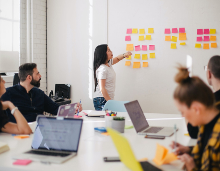 mulher organiza post its na parede, representando uma técnica de agilidade, enquanto outras pessoas do time estão sentadas em uma mesa com seus computadores